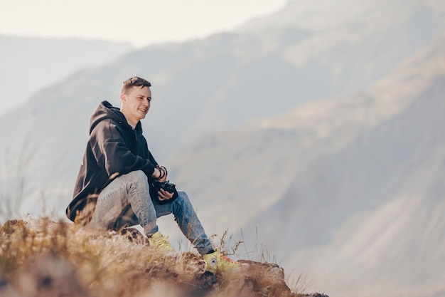 Un joven fotógrafo con una capucha negra sostiene una cámara en sus manos y toma fotografías de una chica posando contra el fondo de las montañas rocosas.