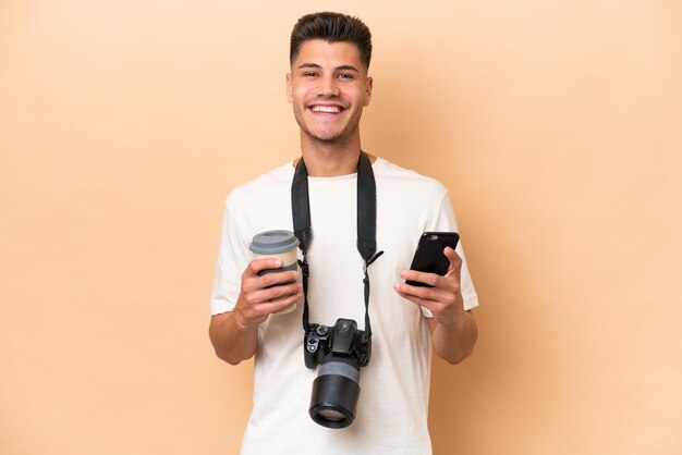 Joven fotógrafo hombre caucásico aislado de fondo beige sosteniendo café para llevar y un móvil