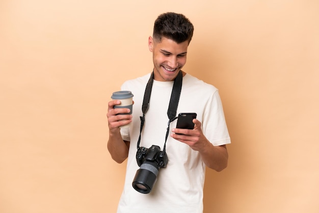 Joven fotógrafo hombre caucásico aislado de fondo beige sosteniendo café para llevar y un móvil