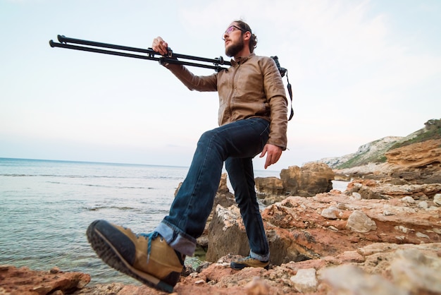 Joven fotógrafo masculino caminando en la playa de roca con cámara en trípode