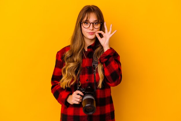 Joven fotógrafo mujer caucásica aislada sobre fondo amarillo con los dedos en los labios guardando un secreto.