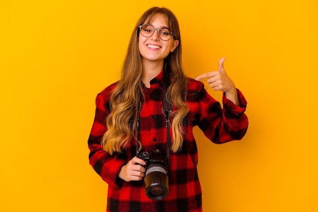 Joven fotógrafo mujer caucásica aislada sobre fondo amarillo persona apuntando con la mano a un espacio de copia de camisa, orgulloso y seguro