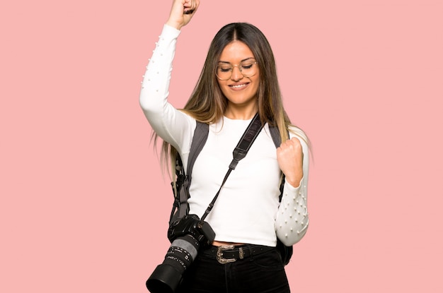 Joven fotógrafo mujer celebrando una victoria en la pared rosa aislada