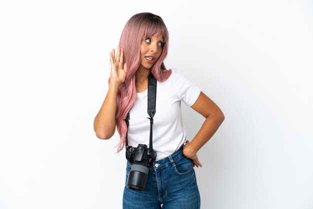 Joven fotógrafo mujer de raza mixta con cabello rosado aislado sobre fondo blanco escuchando algo poniendo la mano en la oreja