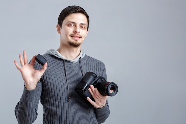 Joven fotógrafo profesional smilling en camisa sostiene la batería para cámara digital DSLR sobre fondo gris