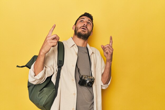 Foto joven fotógrafo de viajes hispano posa apuntando hacia arriba con la boca abierta