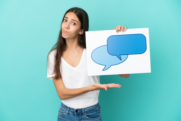 Foto joven francesa aislada de fondo azul sosteniendo un cartel con un icono de burbuja de voz y apuntándolo