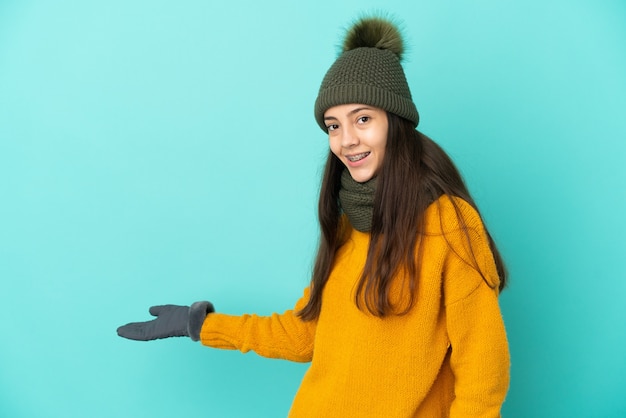 Joven francesa aislada sobre fondo azul con gorro de invierno extendiendo las manos hacia el lado para invitar a venir