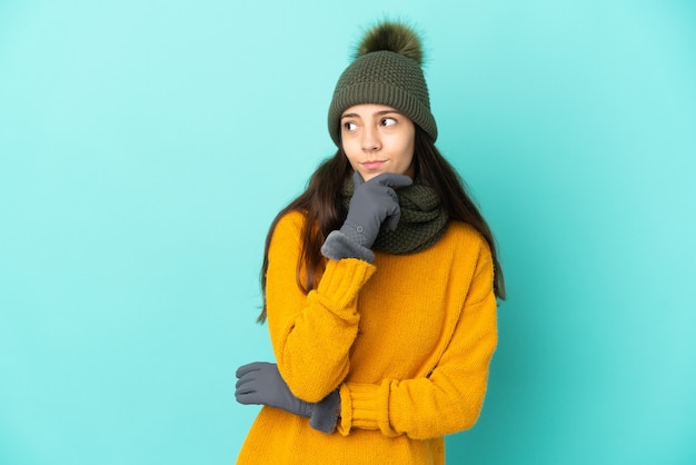 Joven francesa aislada sobre fondo azul con gorro de invierno teniendo dudas