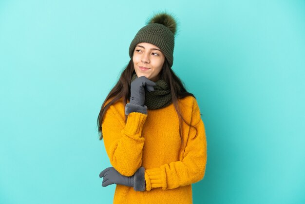 Joven francesa aislada sobre fondo azul con sombrero de invierno y mirando hacia arriba