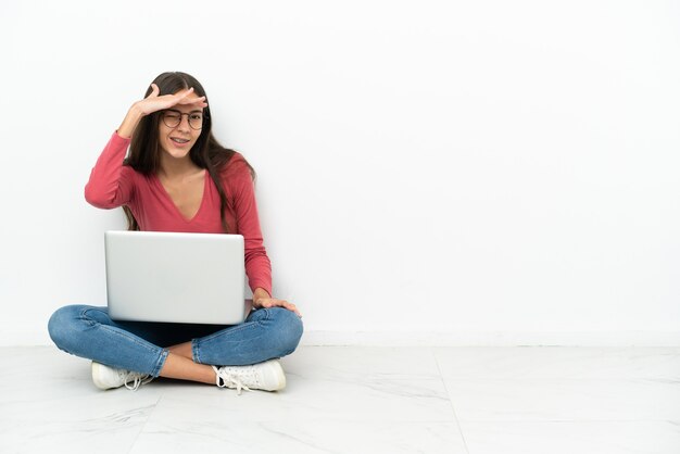 Joven francesa sentada en el suelo con su computadora portátil mirando lejos con la mano para mirar algo