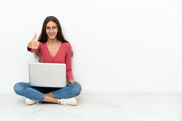 Joven francesa sentada en el suelo con su computadora portátil con los pulgares hacia arriba porque algo bueno ha sucedido