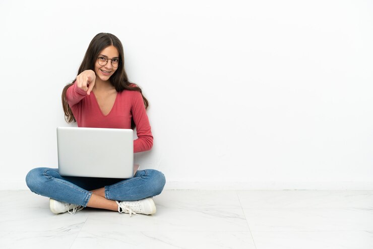 Foto joven francesa sentada en el suelo con su computadora portátil le señala con el dedo con una expresión de confianza