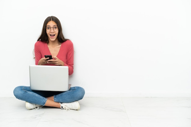 Joven francesa sentada en el suelo con su computadora portátil sorprendida y enviando un mensaje