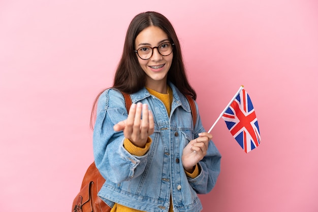 Joven francesa sosteniendo una bandera del Reino Unido aislada sobre fondo rosa invitando a venir con la mano. Feliz de que hayas venido