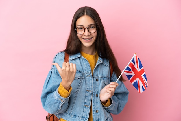 Joven francesa sosteniendo una bandera del Reino Unido aislado sobre fondo rosa apuntando hacia el lado para presentar un producto