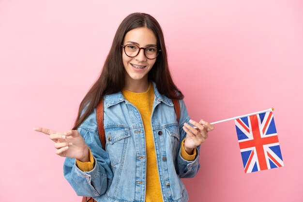 Joven francesa sosteniendo una bandera del Reino Unido aislado sobre fondo rosa dedo acusador hacia el lado