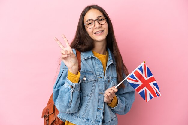 Joven francesa sosteniendo una bandera del Reino Unido aislado sobre fondo rosa sonriendo y mostrando el signo de la victoria