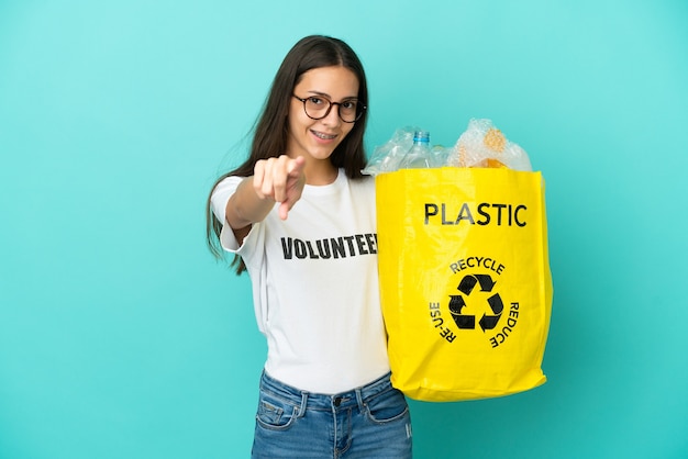 Joven francesa sosteniendo una bolsa llena de botellas de plástico para reciclar apuntando hacia el frente con expresión feliz