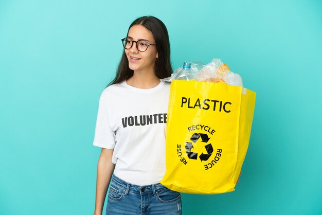Joven francesa sosteniendo una bolsa llena de botellas de plástico para reciclar mirando hacia un lado y sonriendo