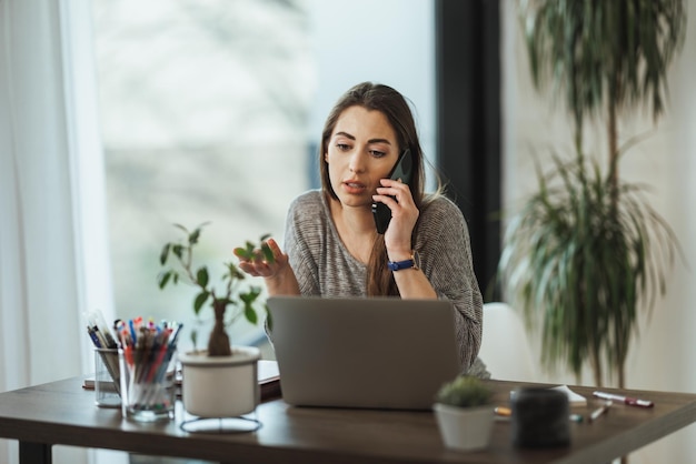 Una joven freelance que se ve reflexiva y habla en un teléfono inteligente mientras trabaja desde su oficina en casa.