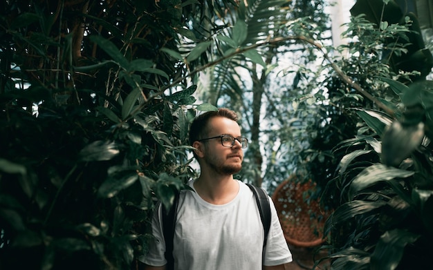 Joven freelancer usa anteojos en el parque tropical Hombre barbudo con camisa blanca mira hacia un lado El tipo de TI usa mochila