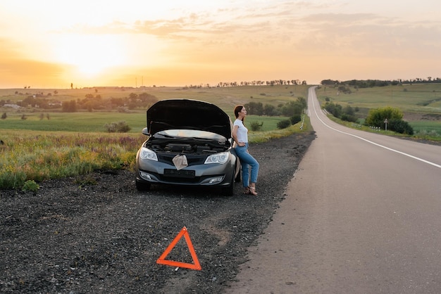 Una joven frustrada se para cerca de un automóvil averiado en medio de la carretera durante la puesta de sol Avería y reparación del automóvil Esperando ayuda Servicio de automóvil Avería del automóvil en la carretera