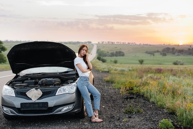 Una joven frustrada se encuentra cerca de un automóvil averiado en medio de la carretera durante la puesta de sol. Avería y reparación del coche. Esperando ayuda.