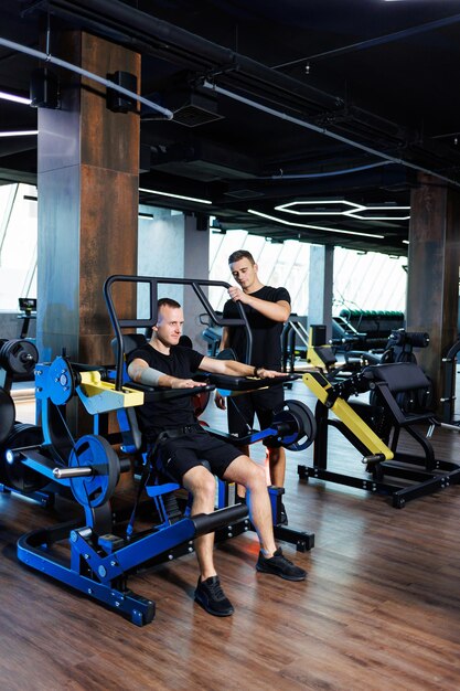 Un joven fuerte realiza ejercicios en el gimnasio bajo la supervisión de un entrenador. Potente entrenamiento realizado por un hombre guapo. Estilo de vida saludable