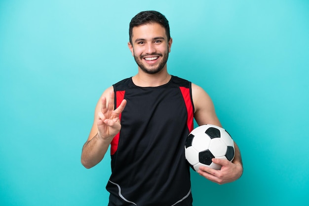 Joven futbolista brasileño aislado de fondo azul feliz y contando tres con los dedos