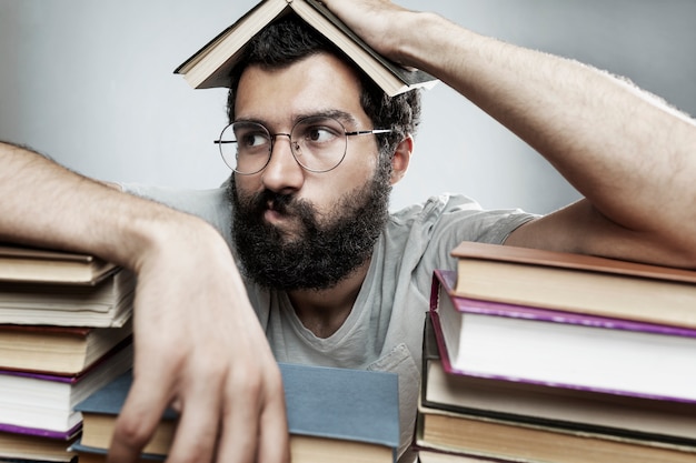 Joven con gafas y una barba se sienta en una mesa con montones de libros. Entrenamiento y educación.