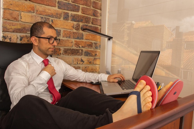 Joven con gafas, camisa, corbata y chanclas mirando la computadora portátil en la oficina en casa