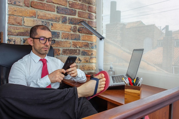 Joven con gafas, camisa, corbata y chanclas sosteniendo un teléfono inteligente en la oficina en casa