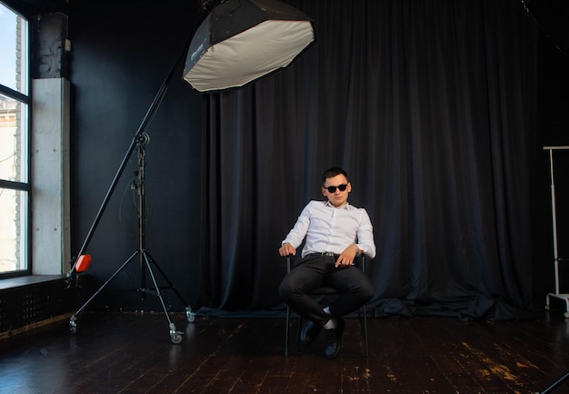 un joven con gafas está sentado en una silla en un estudio fotográfico.