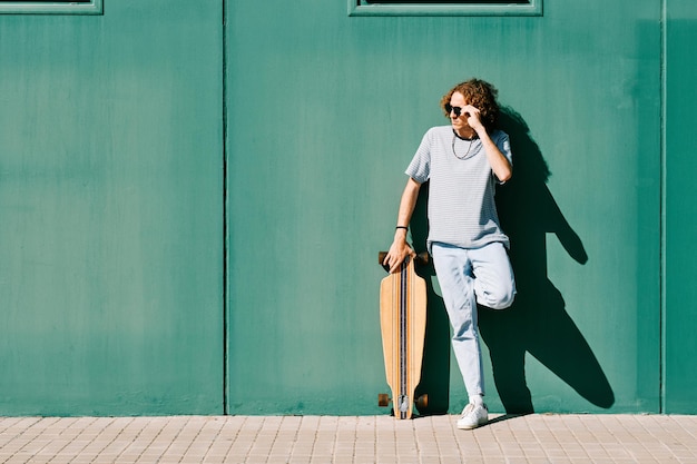 Joven con gafas de sol y longboard o skate en una pared verde en un día de verano