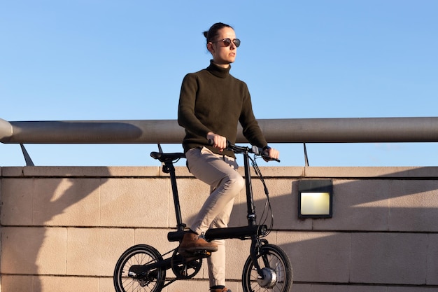 Joven con gafas de sol montando una bicicleta eléctrica por el parque de la ciudad bajo el cielo azul con la luz del atardecer en la cara