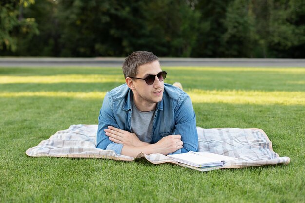 Joven en gafas de sol relajándose y leyendo un libro mientras está acostado en el césped en el parque