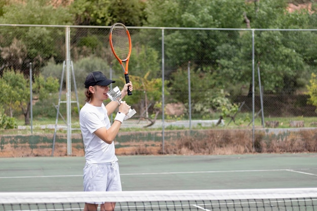 Joven ganando el campeonato de tenis mordiendo la copa como señal de victoria vistiendo traje blanco en una cancha