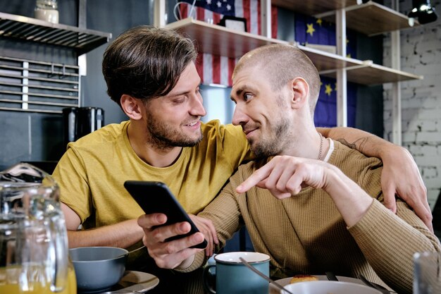 Joven gay abrazando a su novio cuando habla de una nueva aplicación móvil o noticias en las redes sociales durante el desayuno