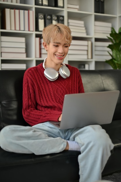 Un joven gay asiático feliz está usando su computadora portátil en un sofá en su sala de estar