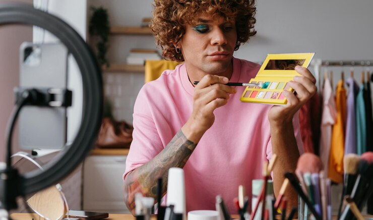 Foto joven gay confiado sosteniendo una paleta de sombra de ojos mientras graba un tutorial de maquillaje desde casa