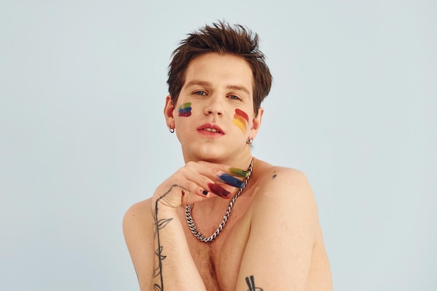 Joven gay está parado en el estudio y posando para una cámara Pintado con maquillaje multicolor