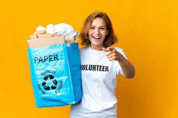 Joven georgiana sosteniendo una bolsa de reciclaje llena de papel para reciclar sorprendida y apuntando hacia el frente