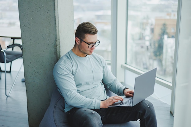 Un joven gerente con anteojos y ropa informal se sienta en una silla suave y trabaja en una computadora portátil Espacio de trabajo para trabajo remoto