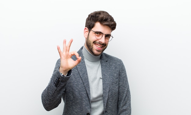 Joven gerente hombre sintiéndose exitoso y satisfecho, sonriendo con la boca abierta, haciendo un signo bien con la pared blanca de la mano