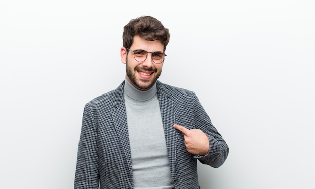 Joven gerente hombre sonriendo alegremente y casualmente, mirando hacia abajo y apuntando al pecho contra la pared blanca