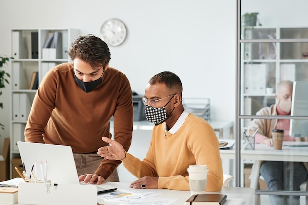 Joven gerente del medio oriente apuntando a la pantalla de la computadora portátil mientras prepara la presentación con un colega durante la pandemia de coronavirus