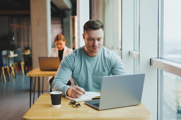 Un joven gerente se sienta en una oficina con grandes ventanales y trabaja en una computadora portátil. Un apuesto hombre de negocios está trabajando en su tarea en un café.