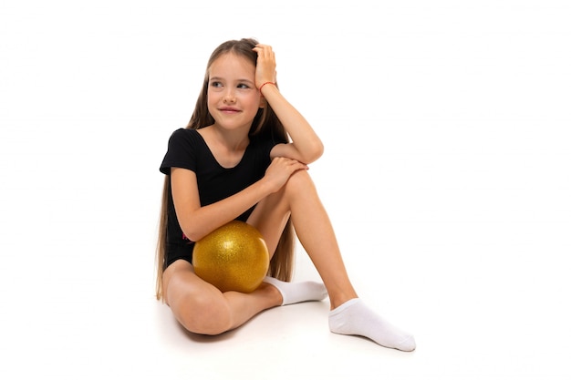 Joven gimnasta niño en un traje de baño deportivo negro se sienta con una pelota sobre un fondo blanco.