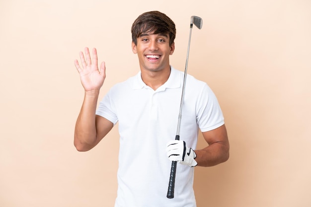 Joven golfista hombre aislado sobre fondo ocre saludando con la mano con expresión feliz